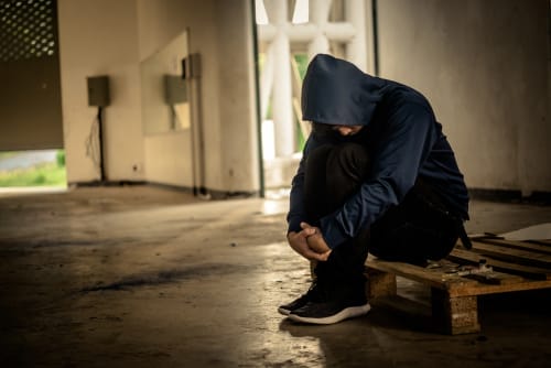 Person in Hoodie Sitting Alone – Opioid Addiction Health Effects - Ferraro Law Firm Person in a hoodie sitting alone in a dark, empty space, symbolizing the physical and emotional toll of opioid addiction.