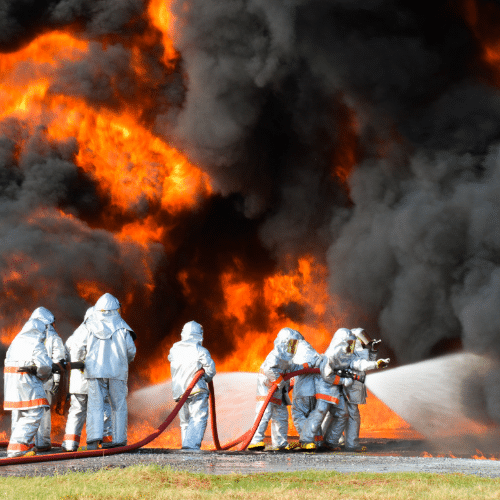 Group of firefighters in silver protective suits spraying AFFF firefighting foam on a massive fire with heavy smoke.