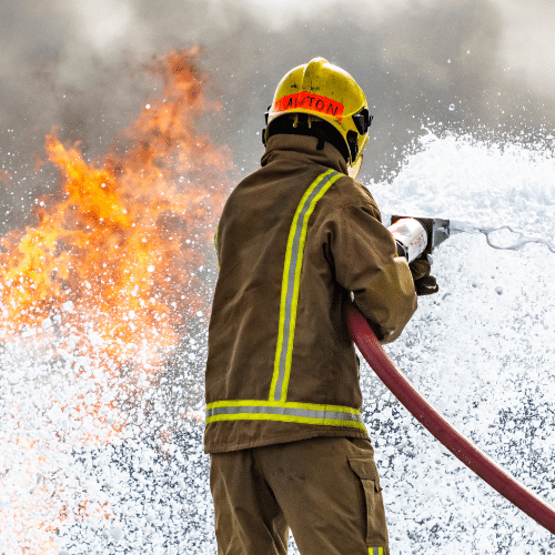 Firefighter in protective gear spraying firefighting foam toward flames, illustrating AFFF exposure risk.