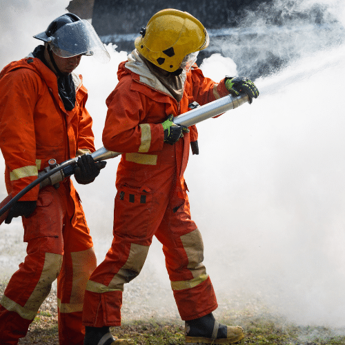Two firefighters in protective gear spraying firefighting foam to control a blaze.