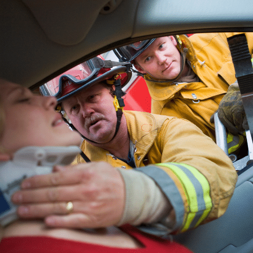 Two firefighters providing emergency assistance to a woman in a car after an accident, stabilizing her neck with a cervical collar.