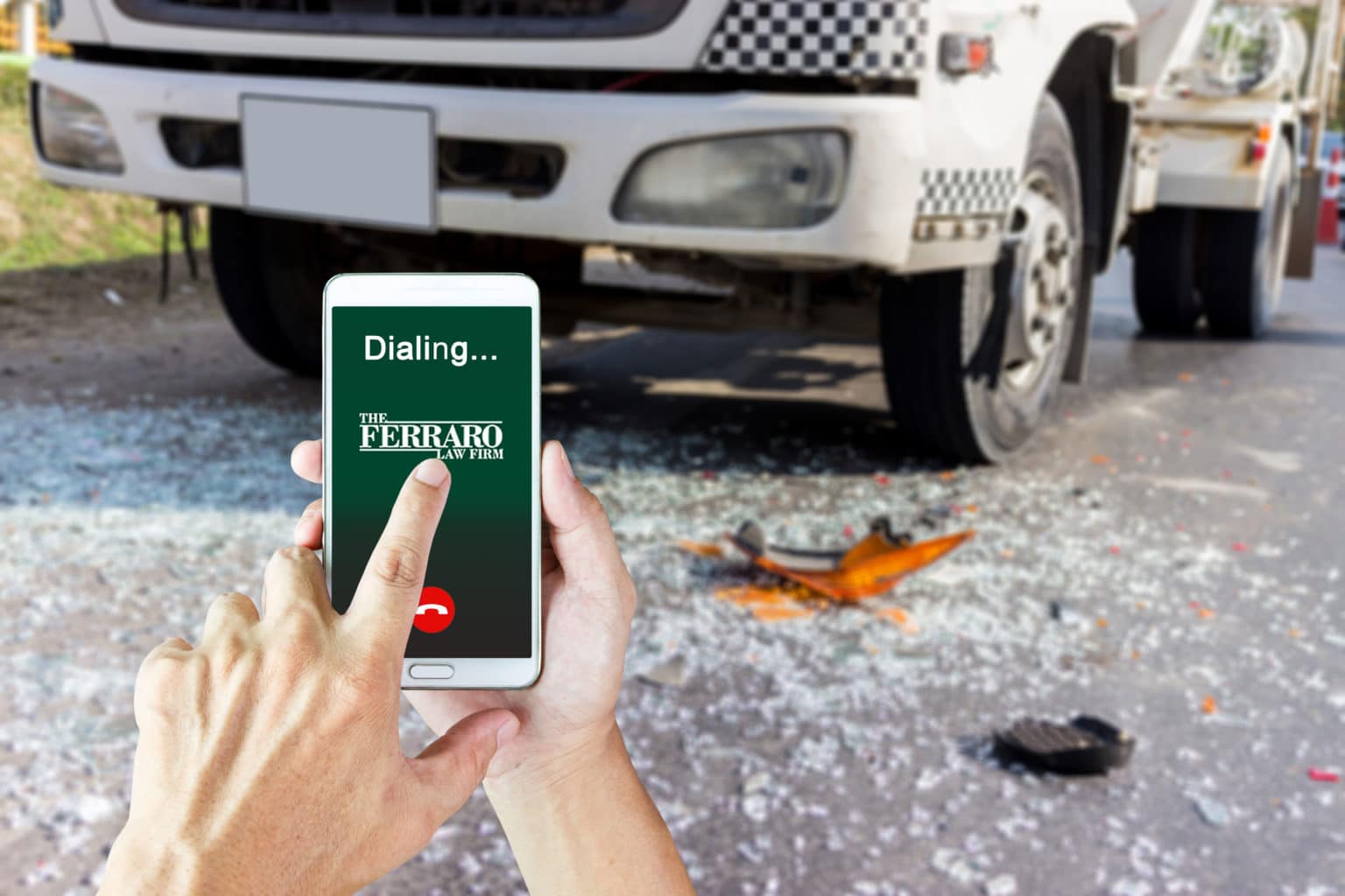 Person holding a smartphone displaying the Ferraro Law logo while dialing, with a damaged semi-truck and shattered glass on the road in the background, symbolizing seeking legal help after a truck accident.