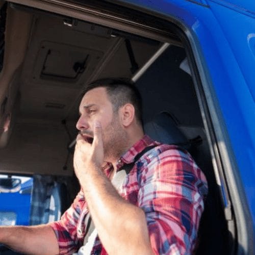 Tired truck driver in a red plaid shirt yawning while sitting in the cab of a blue semi-truck, representing fatigue and accident risks in the trucking industry.