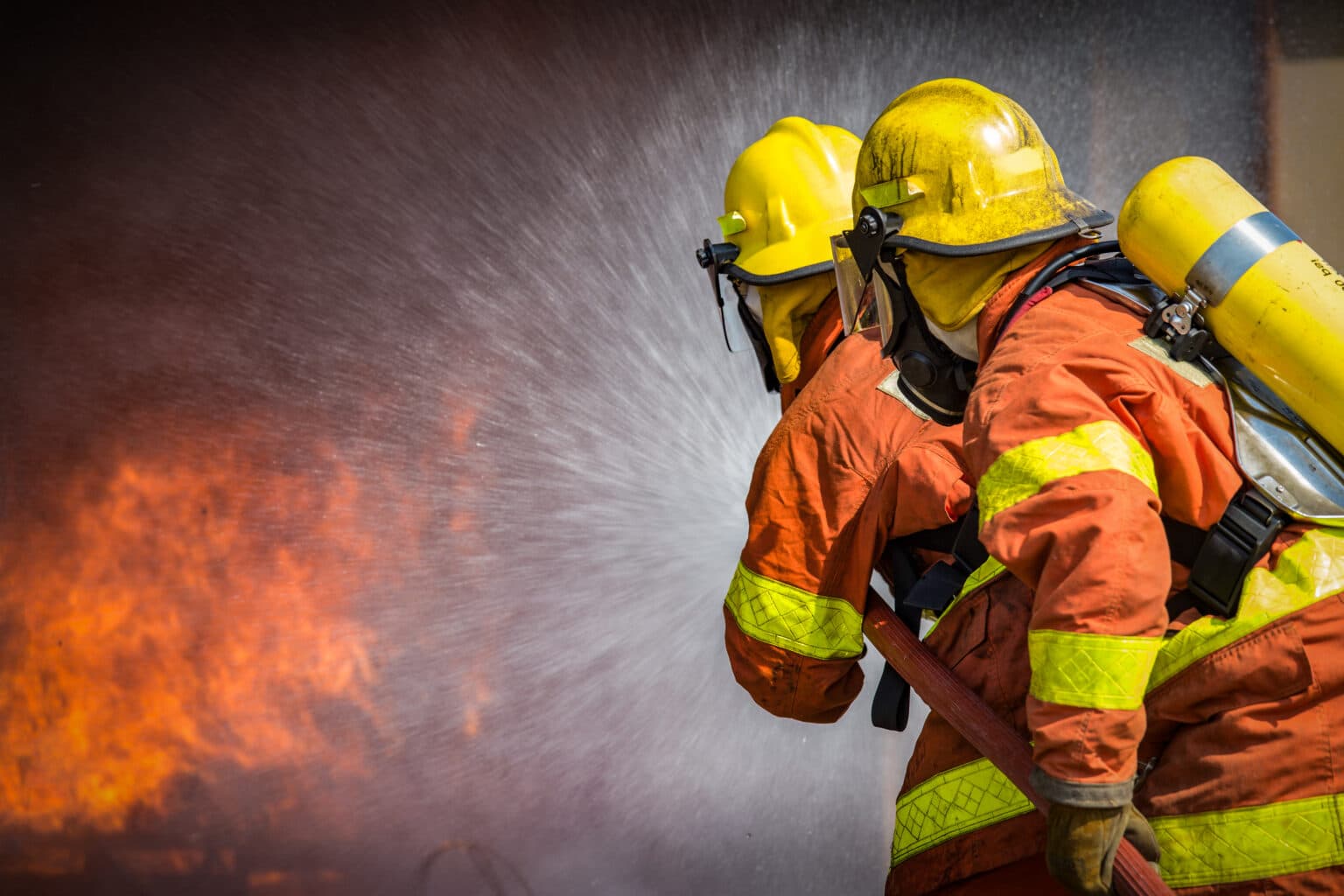 Two firefighters in full protective gear spraying water to control a blaze, illustrating the occupational risks of PFAS exposure from firefighting foam and equipment.