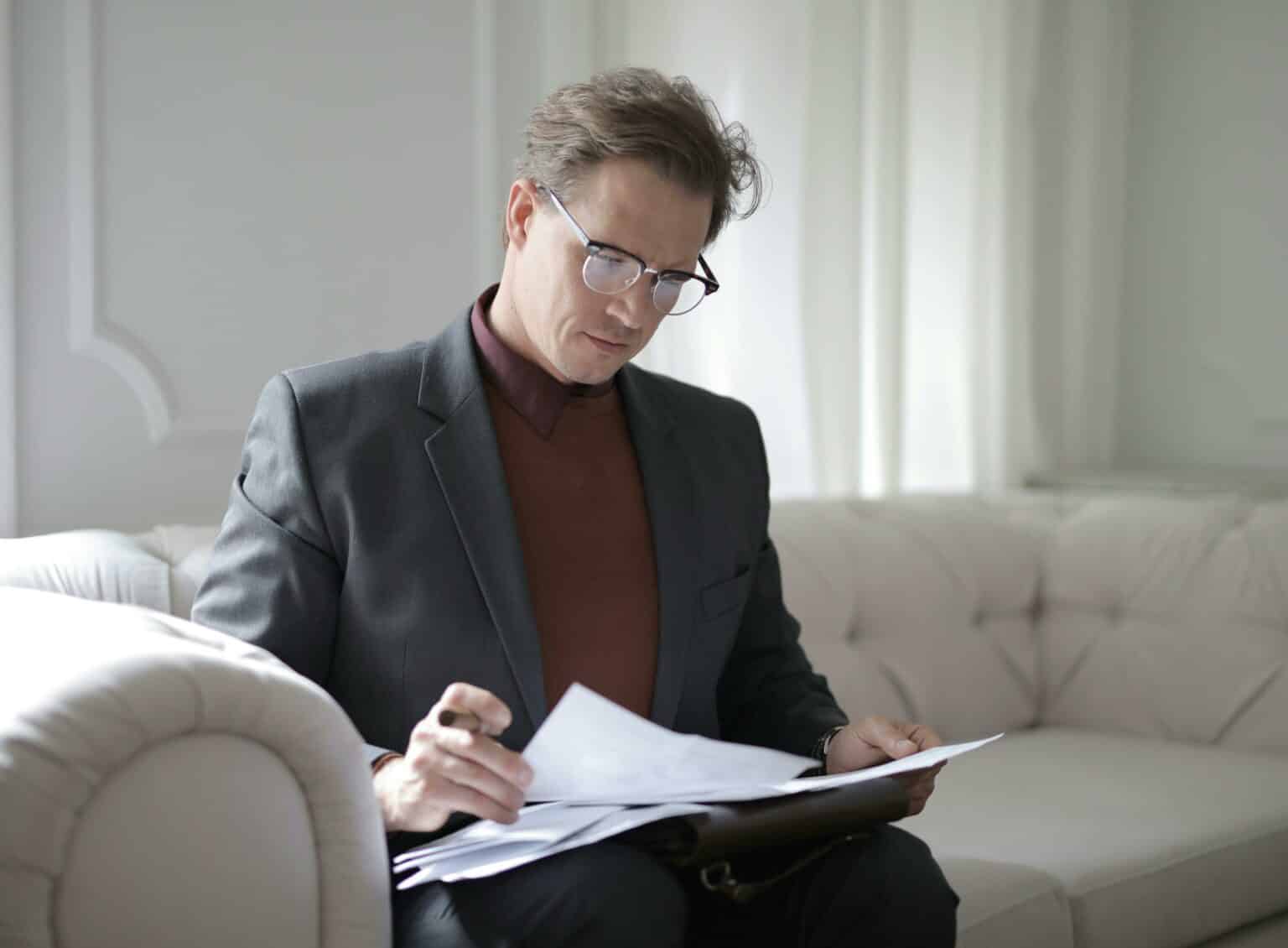 Man in a suit and glasses reviewing legal documents on a sofa, representing a client meeting with a wrongful death attorney to prepare questions for their case. Man in a suit and glasses reviewing legal documents on a sofa, representing a client meeting with a wrongful death attorney to prepare questions for their case.